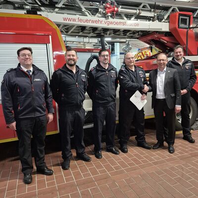 Sechs M&auml;nner stehen vor einem Feuerwehrauto. Einer h&auml;lt eine Urkunde fest. Er hat eine neue Aufgabe bekommen.