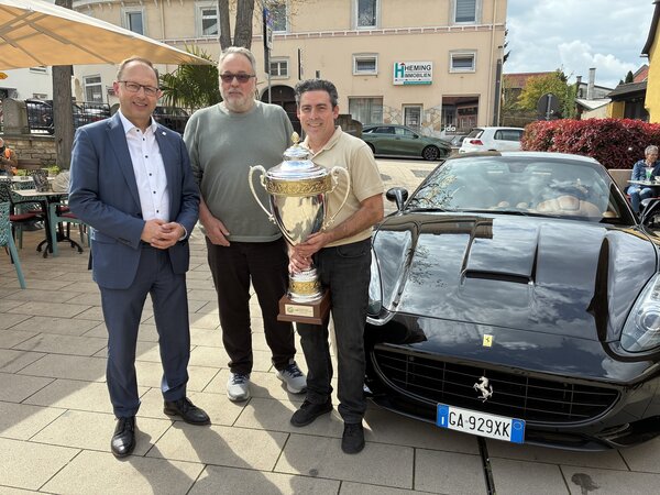 Drei M&auml;nner stehen mit einem Pokal vor einem Auto. 