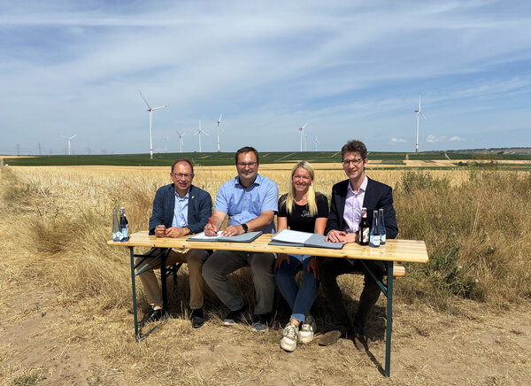 Das Bild zeigt vier Personen auf einer Bank sitzend vor einem Getreidefeld. Sie unterzeichnen einen Vertrag.
