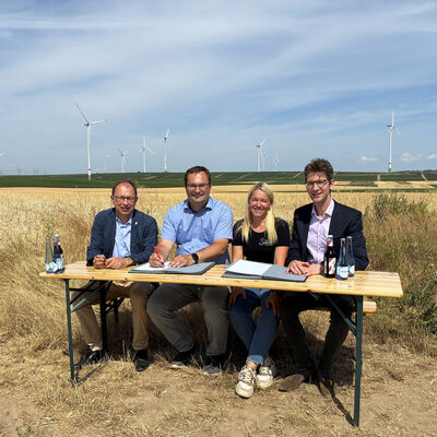 Das Bild zeigt vier Personen auf einer Bank sitzend vor einem Getreidefeld. Sie unterzeichnen einen Vertrag.