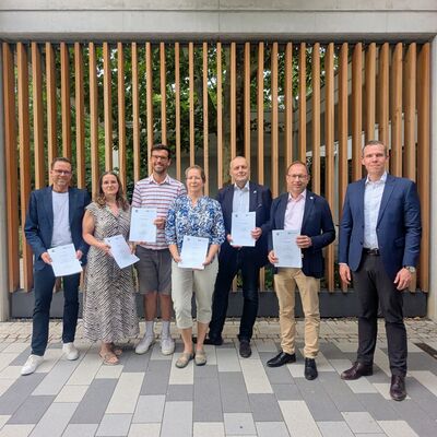 Das Bild zeigt eine Gruppe von Personen f&uuml;r ein Foto aufgereiht. Sie halten vertragsunterlagen in ihren H&auml;nden. 