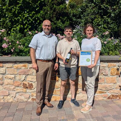 Das Bild zeigt drei Personen vor einer Natursteinmauer. Sie halten den pr&auml;mierten Weinfestwein und die Urkunde in den H&auml;nden. 